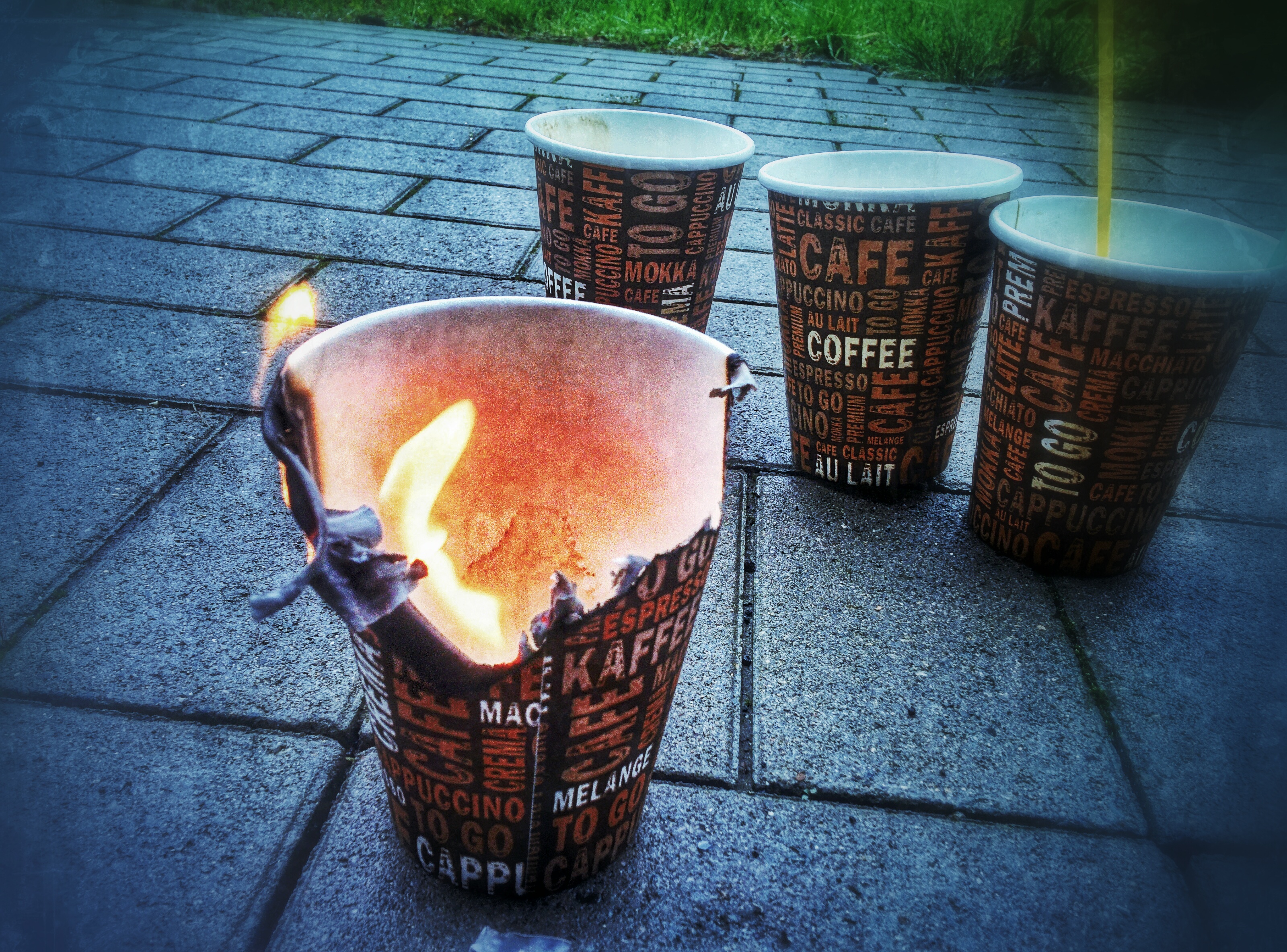 Image of 4 paper coffee mugs standing on paving bricks. One of them is separated from the others and on fire.